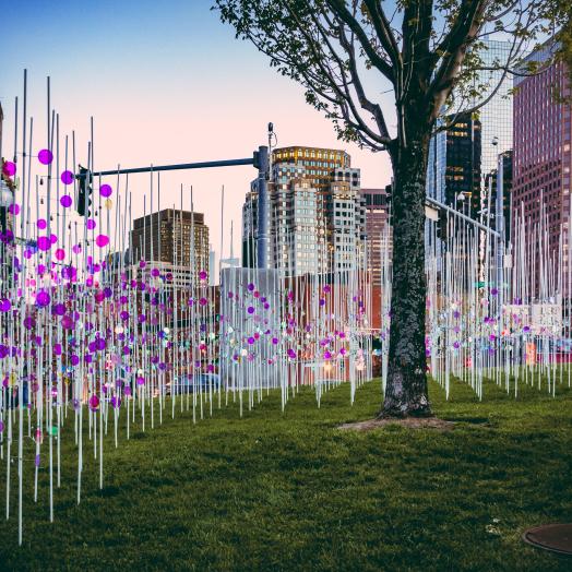 A large array of aluminum poles with fixed circles of colored glass, together forming a wave pattern, stand in a green field with some tall buildings in the background.